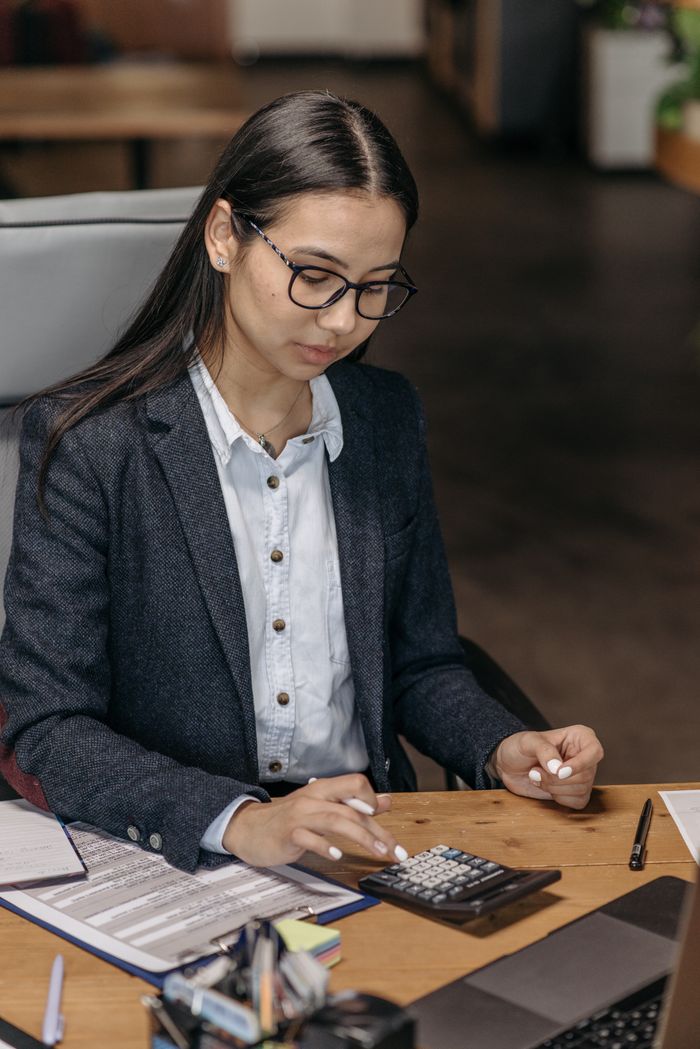 Home Professional Asian woman in a blazer using calculator at desk, calculating finances.